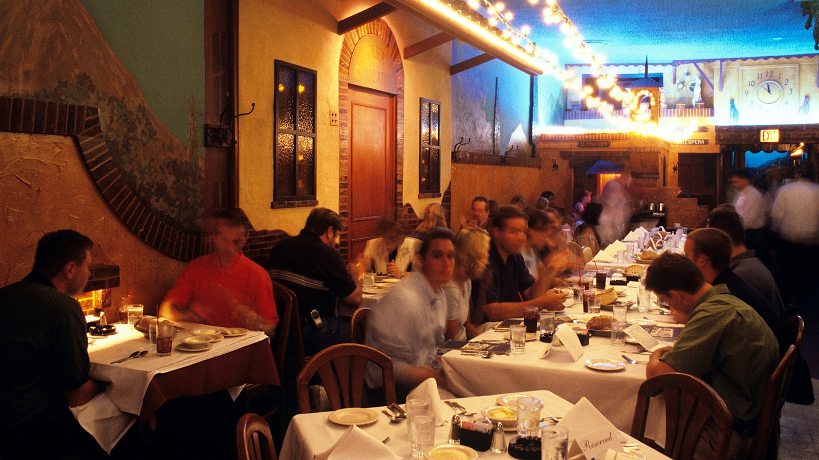 People eat in the dining room of the Village, with lights hanging from above, blue walls and a wall designed to look like the exterior of a traditional house in Italy